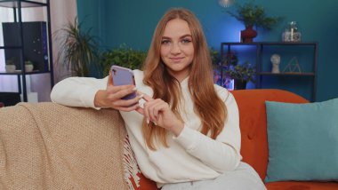 Portrait of adult girl sitting on sofa uses mobile phone smile at modern home apartment. Young woman texting share messages content on smartphone social media applications online, watching relax movie