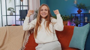 Happy caucasian girl shouting, celebrating success, winning, goal achievement, good news, raising fists in gesture I did it, lottery luck at home apartment. Young woman indoors in living room on couch