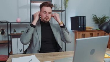 Happy relaxed overjoyed businessman in suit working on laptop computer at office wearing earphones listening favorite energetic disco music on smartphone. Male freelancer man relaxing, taking a break