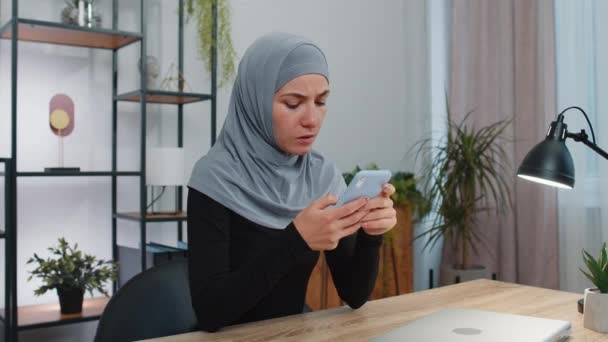 ヒジャーブスカーフの若いイスラム教徒のビジネス女性は スマートフォンの入力ブラウジングを使用し 突然の宝くじの結果 悪いニュース 財産の損失 失敗に驚いて失う 現代のホームオフィスワークスペースの女の子 — ストック動画