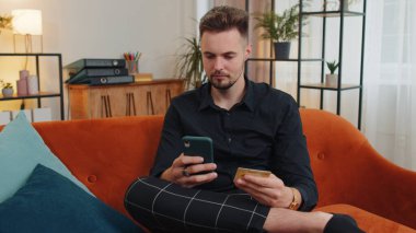Modern ev apartmanında oturan ve çevrimiçi yemek siparişi veren, kredi kartı ile ödeme yapan mutlu tüketici genç adam. İnternette indirimli satış. Kanepede yetişkin bir adam.