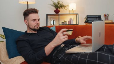 Para transfer ederken kredi kartı ve dizüstü bilgisayar kullanan, online alışveriş yapan, eve yemek siparişi veren bir adam. Oturma odasında elinde dolarla kanepede oturan genç adam.