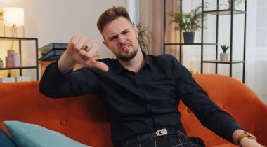 Dislike. Upset man showing thumbs down sign gesture, expressing discontent, disapproval, dissatisfied bad work at modern home apartment indoors. Displeased young guy in living room sitting on sofa