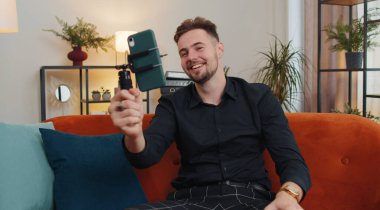 Yakışıklı erkek blogcu cep telefonu kamerasından selfie çeken cihaz ekranına bakıyor, modern sosyal medya vlog uygulamasında sanal video görüşmesi yapıyor. Evdeki koltukta akıllı telefonu olan adam.