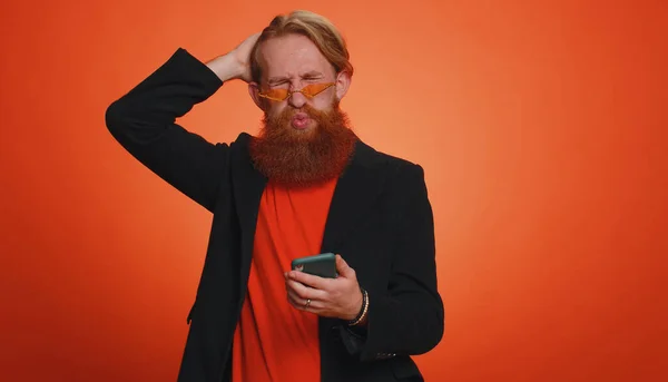 ジャケットのひげを生やした男は 携帯電話の入力ブラウジングを使用して 突然宝くじの結果 失敗に驚いて失う 若いです赤毛男男の子絶縁一人でオレンジ色のスタジオ背景 — ストック写真