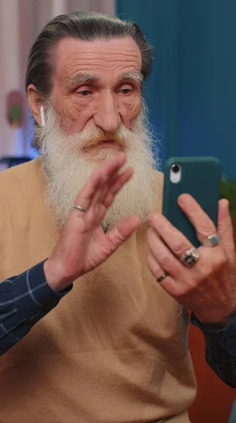 高齢の祖父は ソファの上の自宅のリビングルームで家族とスマートフォンのオンライン会話を呼び出す作るイヤフォンを身に着けている男 引退したおじいちゃん年金受給者モバイルスピーカーの話を楽しんで — ストック写真