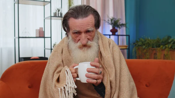 Sick senior grandfather man wrapped in plaid shivering from cold on sofa drinking hot tea in unheated apartment without heating due debt. Unhealthy elderly grandpa feeling discomfort try to warming up