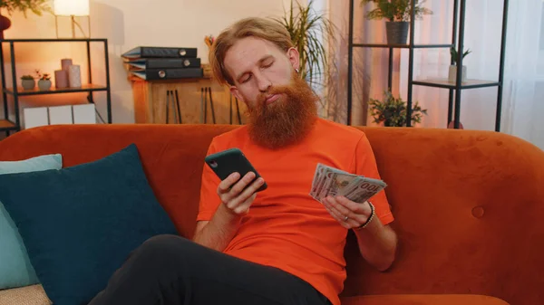 规划家庭预算 满头胡须的红头发的男人用智能手机计算家里的账单 对收入感到满意的快乐的年轻人为计划的假期 礼物存钱 — 图库照片