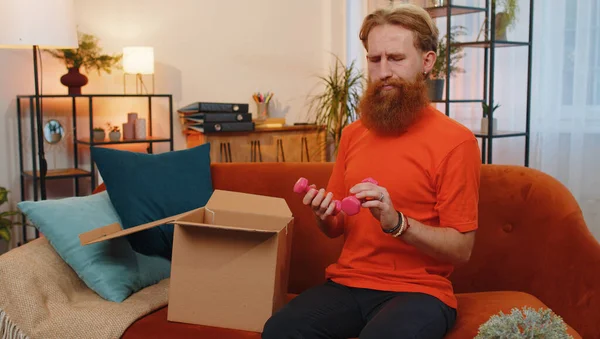 Angry dissatisfied shopper bearded man unpacking parcel feeling upset and confused with the wrong dumbbells delivery from an online sport store, bad quality purchase. Young guy at home on orange couch