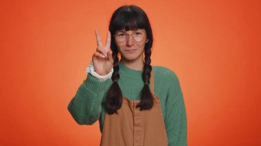 Yeşil kazaklı hippi kadın zafer işareti gösteriyor, başarı ve kazanma umuduyla barış jesti yapıyor, iyimser bir ifadeyle gülümsüyor. Portakal rengi stüdyoda izole edilmiş genç bir kız.