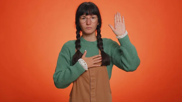 I swear to be honest. Sincere responsible young nerd woman raising hand to take oath, promising to be honest and to tell truth, keeping hand on chest. Latin girl isolated on studio orange background