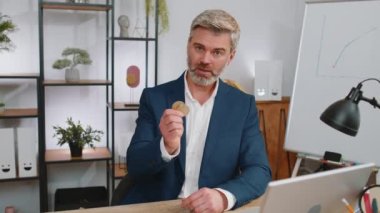 Olgun bir borsa simsarı çevrimiçi ticaret operasyonlarını izledikten sonra bitcoin kazanıyor. Zenginliği artırmak, finansal refahı arttırmak. Serbest çalışan orta yaşlı bir adam iş yerinde kameraya konuşuyor.