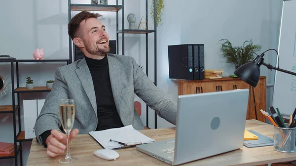 Businessman working on laptop celebrate successful contract agreement with colleague client at office workplace desk. Freelancer man drinking champagne. Online remote video call conversation job