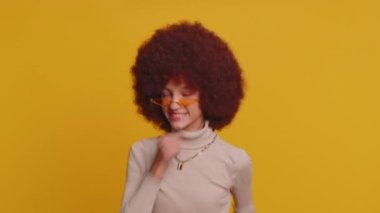 Modayı takip eden neşeli, afro saç stili olan, neşeli, genç bir kız. Dans edip ritme ayak uyduran, el kaldıran, dublaj yapan. Sarı arka planda genç bayan çocuk.