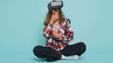 Genç kız çocuk sanal gerçeklik teknolojisi VR uygulama kulaklığı kullanarak 3D video oyunu simülasyonu yapıyor, film izliyor. Gözlüklü çocuklar mavi arka planda otururlar.