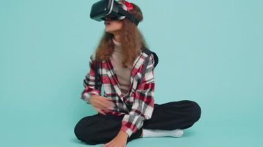 Genç kız çocuk sanal gerçeklik teknolojisi VR uygulama kulaklığı kullanarak 3D video oyunu simülasyonu yapıyor, film izliyor. Gözlüklü çocuklar mavi arka planda otururlar.