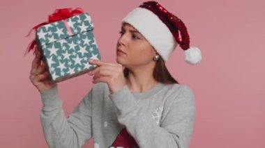 Noel Baba süveteri giymiş kafası karışmış genç bir kadın Noel tatilini kutluyor. Sarılmış Noel hediyesi paketini sallıyor, kulağına yakın duruyor ve tahmin et içinde ne var. Pembe arka plandaki kız