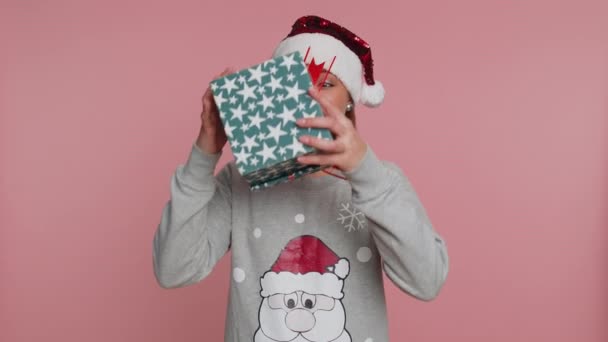 サンタ 句のセーターの若い女性はクリスマス休暇を祝いますギフトボックスを受け取り 素敵なプレゼント 予期しない驚きに満足して見て楽しく笑っています 女の子オンピンクスタジオの背景 — ストック動画