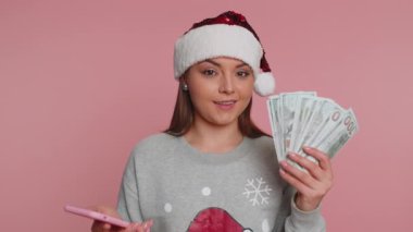 Noel Baba 'nın Noel Baba süveteri içinde mutlu bir kadın. Akıllı telefon görünümlü. Sevinçli bir şekilde kazanan, nakit para alan, piyango talihi kazanan. Pembe arka planda izole edilmiş genç kız