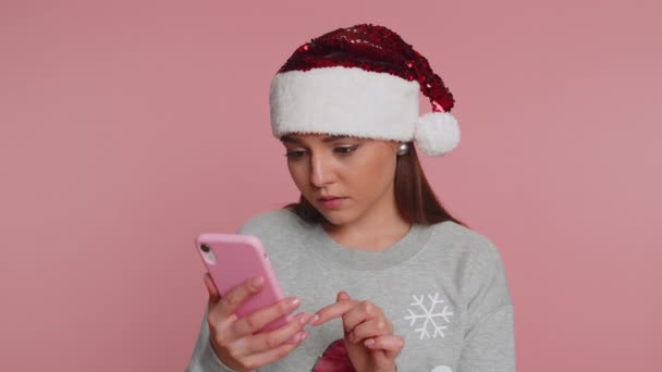 クリスマスサンタ句セーターの素敵な女性は 携帯電話の携帯電話の入力のブラウジングを使用し 突然の宝くじの結果 失敗に驚いて失う 若いです女の子オンピンクスタジオ背景 — ストック動画