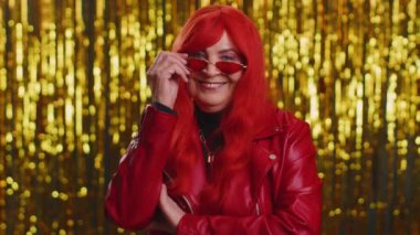 Oyuncu, mutlu, stil sahibi, 70 yaşında, sarı güneş gözlüğü takmış, kameraya bakan, dişlek gülümsemeli, göz kırpan ve iyimser bir şekilde flört eden yaşlı bir kadın. Parlak duvarda kıdemli büyükanne