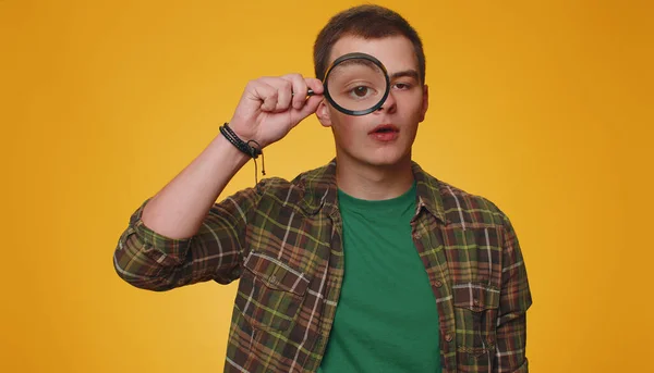 Investigator researcher scientist man holding magnifying glass near face, looking into camera with big zoomed funny eyes, searching, analysing. Young adult guy boy isolated on yellow studio background