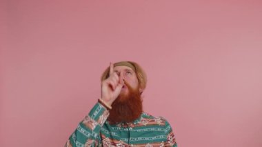 Hippi kızıl sakallı, desenli tişörtlü, başparmağını kaldırıp boş alanı işaret eden bir adam. Reklam logonuz, afişiniz, mesajınız için yer açın. Pembe arka planda hippi kızıl adam.