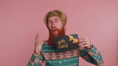 Genç hippi kızıl adam hediye indirimli sertifika kuponuna işaret ediyor. Hipster kızıl saçlı, alışveriş yılbaşı sürpriz kartı olan, pembe arka planda izole edilmiş.