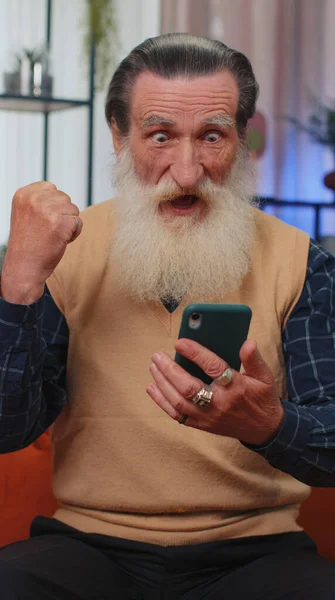 幸せな興奮の高齢者の祖父の男性は 携帯電話のスマートフォンのタイピングブラウジングを使用するすごいはい宝くじのジャックポットを祝う素晴らしい勝利良いニュースが判明したと言う 定年退職したおじいちゃん年金受給者 垂直表示 — ストック写真