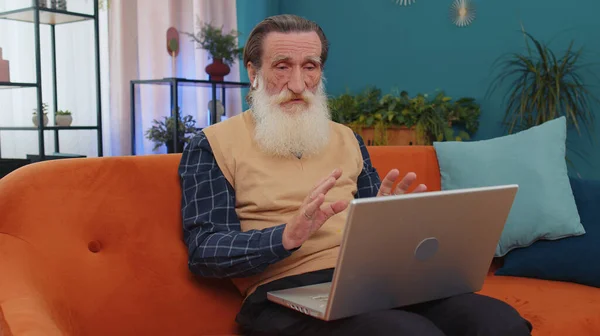 Senior old grandfather sitting on couch, looking at laptop, making video webcam conference call with friends or family, enjoying pleasant conversation. Elderly man laughing, waving hello alone in home