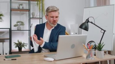 Çatışma, tartışma, pazarlık. Öfkeli olgun iş adamı ofis masasında webcam laptop çevrimiçi video görüşmesi yapıyor. Kızgın, serbest çalışan, kır saçlı, yaşlı bir adam iş arkadaşlarıyla içeride konuşuyor.