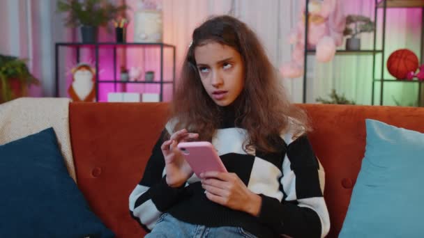 ティーンの女の子のテキストメッセージは リラックスした映画 ニュースを見てオンラインスマートフォンのソーシャルメディアアプリケーション上のメッセージコンテンツを共有します 若いです 子供家の中で夜のリビングプレイルームオレンジのソファに座って — ストック動画