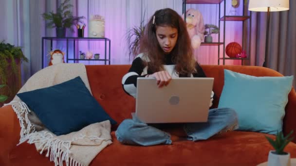 ソファに座って自宅のナイトプレイルームで素敵な十代の女の子のフリーランサーは ラップトップが動作を開始開きます 子供はノートブックで働き メッセージを送ったり オンラインで購入したり 映画を見たり 作業したりします — ストック動画