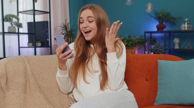 Portrait of lovely girl use mobile smartphone typing browsing say Wow yes found out great big win good news celebrate doing winner gesture. Young woman at home in living room sitting on orange sofa