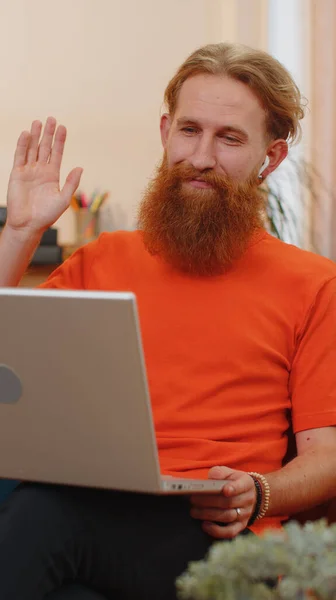 Portrait of bearded redhead man sitting on couch, looking at camera, making video webcam conference call with friends or family enjoying pleasant conversation. Young guy laughing, waving hello at home
