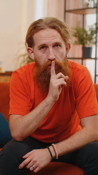 Shh be quiet, hush please. Adult bearded man presses index finger to lips makes silence hush gesture sign do not tells gossip secret. Young guy at home apartment living room sitting on orange couch