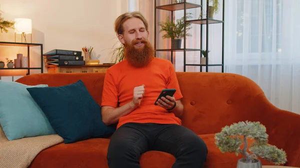 Portrait of bearded man sitting on sofa uses mobile phone smiles at modern home apartment. Young guy texting share messages content on smartphone social media applications online, watching relax movie