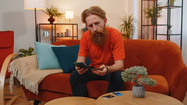 有胡子的男子在汇款 网上购物 在家里订购食物时使用信用卡和智能手机 客厅里坐在沙发上的红头发小伙子 — 图库照片