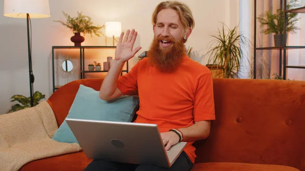 Portrait of bearded redhead man sitting on couch, looking at camera, making video webcam conference call with friends or family, enjoying pleasant conversation. Young guy laughing waving hello at home