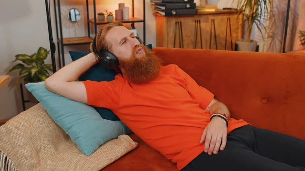 自宅でお気に入りのエネルギッシュなディスコダンスミュージックを聴きながら オレンジのソファの上に寝そべってリラックスしているワイヤレスヘッドフォンの幸せな髭の若者 週末のレジャー活動 ライフスタイル — ストック写真