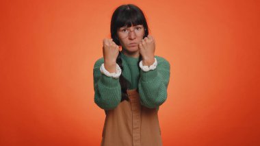 Saldırgan milenyum kadını kameraya savaş tehdidi savuruyor, yumruk tokalaştırıyor, ifadeyle boks yapıyor, cezalandırıyor, negatif öfke egzersizleri yapıyor. Genç kız turuncu arka planda tek başına.