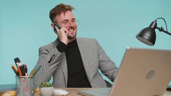 オフィスのワークスペースのデスクに座っている幸せな髭ビジネスマンの上司は 電話で会話をする 若いですフリーランサー男男の子楽しみますモバイル話同僚とともにブルースタジオの背景 — ストック写真