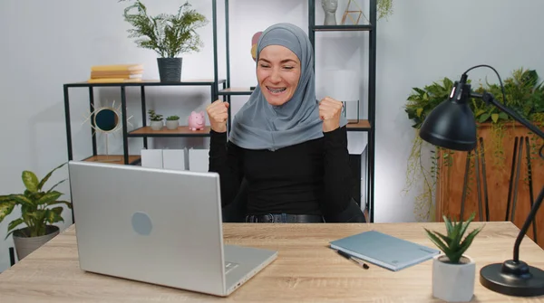 Concentrated business woman winner in hijab working on laptop at home office desk typing browsing say Wow yes found out great big lottery win good news celebrate. Muslim freelancer girl occupation