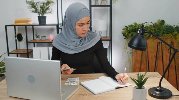 Rich business woman in hijab working on laptop pc counting money cash, calculate earnings income profit at office workplace. Professional manager freelancer girl. Muslim people. Employment, occupation
