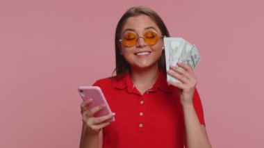 Akıllı telefon görünümlü mutlu turist kadın içten bir sevinç gösterisi kazan, para dolar banknotları al, başarı piyango şansı. Genç milenyum kızı pembe stüdyo geçmişinde tek başına izole edilmiş.