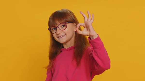 年轻的学龄小女孩戴着眼镜 赞许地看着摄像机 展示出很好的手势 就像积极的手势 认可一些好的东西 在工作室黄色背景下被隔离的未成年女童 — 图库照片