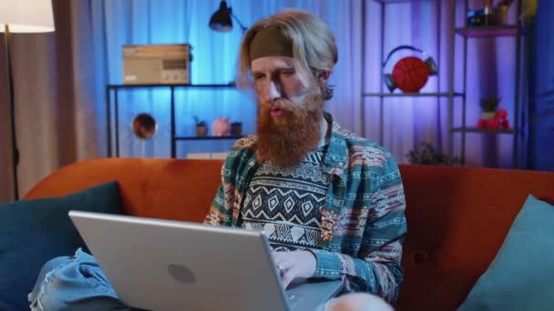 自宅のソファでリビングルームで仕事を終えた後 ソファの上に座って髭を生やした男 若い男のフリーランスは オンラインで働いてリモートジョブ 電子学習 ノートPc上でインターネットを閲覧 — ストック動画