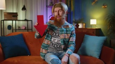 Evdeki koltukta oturan sakallı sakallı hippi adam kırmızı oku gösteriyor. Düşük not kavramı, başarısız iş anlayışı, borsa döviz kurunun düşüşü iflas. Gece odasındaki hippi.