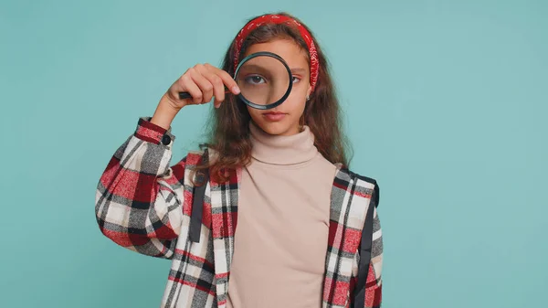 Investigator researcher scientist teenager child girl kid holding magnifying glass near face, looking into camera with big zoomed funny eye, searching, analysing. Preteen children on blue background