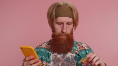 Samimi kızıl saçlı hippi müşteri kredi kartı ve akıllı telefon kullanarak online alışveriş ve ödeme yapıyor. Finans ve internet. Genç adam pembe arka planda tek başına.